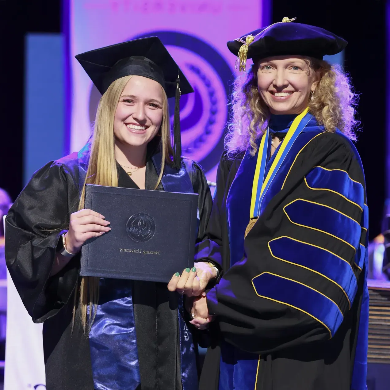 A Herzing University graduate smiling as she receives her diploma on stage, alongside a faculty member in ceremonial regalia during commencement.